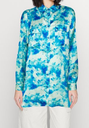 Chemise à manches longues boutonnée avec motif tie-dye bleu et vert, deux poches poitrine avec boutons, portée avec un pantalon blanc.