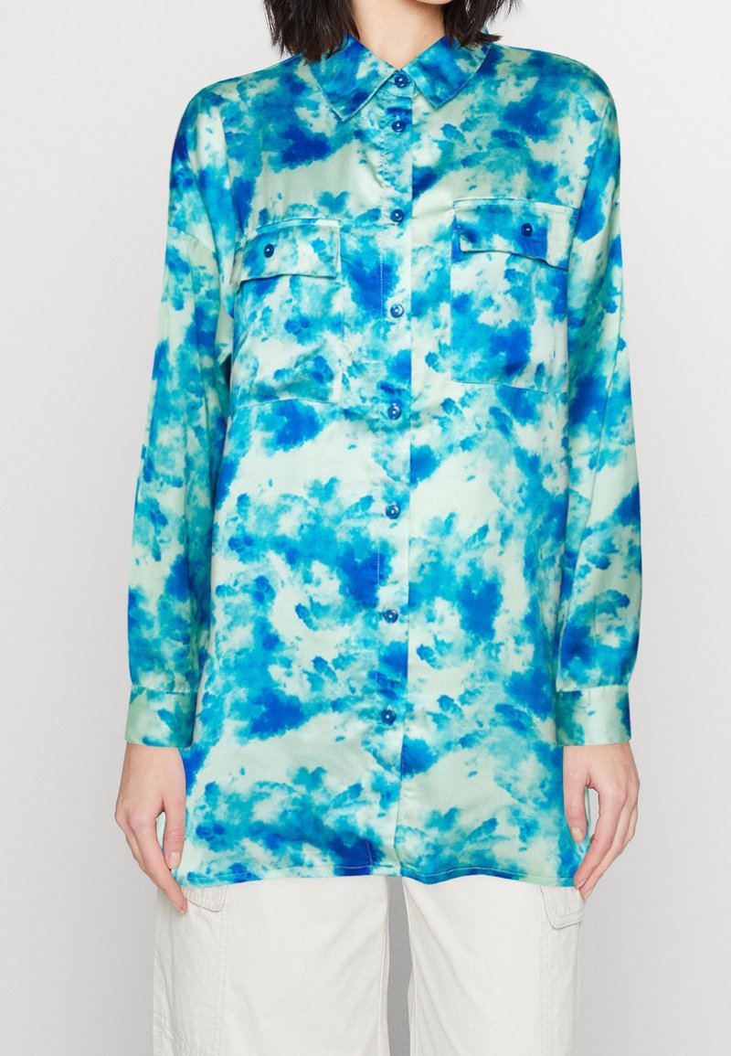 Chemise à manches longues boutonnée avec motif tie-dye bleu et vert, deux poches poitrine avec boutons, portée avec un pantalon blanc.