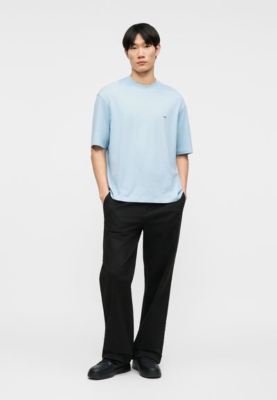 Άνδρας που στέκεται με τα χέρια στις τσέπες, φορώντας ανοιχτό μπλε πουκάμισο με κοντά μανίκια, μαύρο παντελόνι και μαύρα παπούτσια, μπροστά σε απλό φόντο.