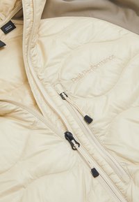 Giacca puffer beige con design trapuntato, dotata di un colletto alto, due zip e il logo "PeakPerformance" ricamato in filo dorato.