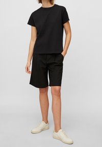 Persona con una camiseta negra de manga corta, pantalones cortos negros hasta la rodilla y zapatillas blancas de pie contra un fondo claro y liso.