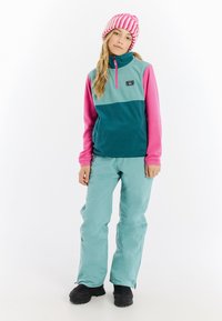 Fleece pullover in teal en roze met een halve rits, gecombineerd met lichtteal broeken en zwarte laarzen. Bevat een logo op de borst.