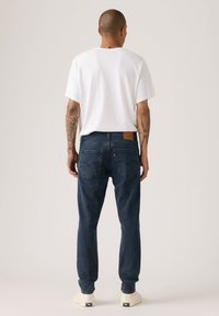 Tamsiai mėlynos džinsinės kelnės, tiesaus kirpimo, vidutinio svorio džinsas, su rudos odos aplikacija ir išskirtinėmis galinėmis kišenėmis. Derinamos su balta marškyte.