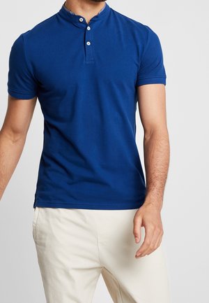 Mann trägt ein eng anliegendes blaues Henley-Shirt mit kurzen Ärmeln, weißen Knöpfen und hellbeiger Hose vor einfarbigem Hintergrund.