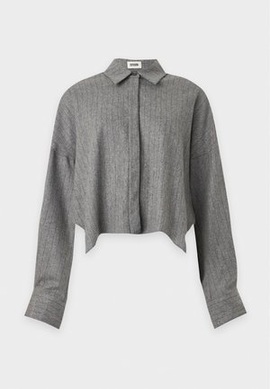 Une femme porte une veste grise, un col roulé marron, une jupe noire et des bottes hautes jusqu’aux genoux. Elle tient un grand cabas brun olive sur fond blanc.