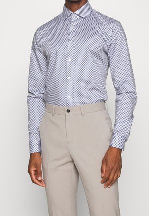 Mann trägt ein langärmeliges Hemd mit geometrischem Muster und hellbeige maßgeschneiderte Hosen vor einfachem Hintergrund.