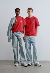 T-shirts vermelhas de algodão com um logótipo, combinadas com jeans de ganga azul desbotados. Um modelo segura um casaco de ganga cinza claro, posando sobre um fundo neutro.