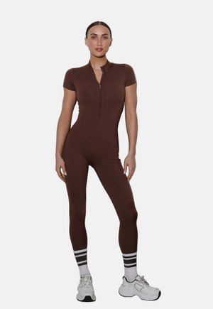 Kobieta stojąca, ubrana w dopasowany brązowy kombinezon z krótkim rękawem i zamkiem z przodu, białe skarpetki w paski oraz białe trampki na jednolitym tle.