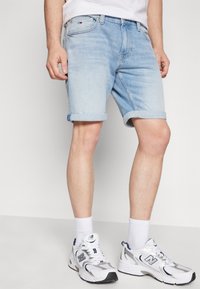 Pantalones cortos de mezclilla azul claro con dobladillos enrollados, cinco bolsillos y cierre de cremallera. Llevados con zapatillas deportivas blancas y calcetines blancos.