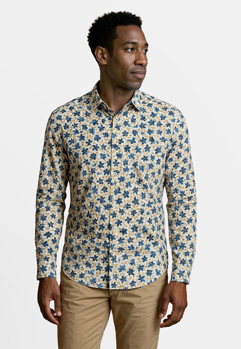 Man in beige broek draagt een knoopbloemige shirt met blauwe en beige bloemen, kijkt naar rechts tegen een effen achtergrond.