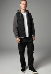 Czarna pikowana kamizelka na szarym hoodie, zestawiona z czarnymi spodniami cargo. Noszona z czarnymi sneakersami z białą podeszwą.
