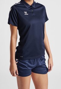 Námořnické polos tričko s krátkými rukávy a límečkem, s bílým logem a designem na rameni. V kombinaci s odpovídajícími námořnickými sportovními šortkami.