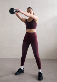 Idrettsutøver i en kastanjerød sports-BH og matchende tights, som utfører en kettlebell-sving. Sorte joggesko og hvite ankelstrømper fullfører antrekket.