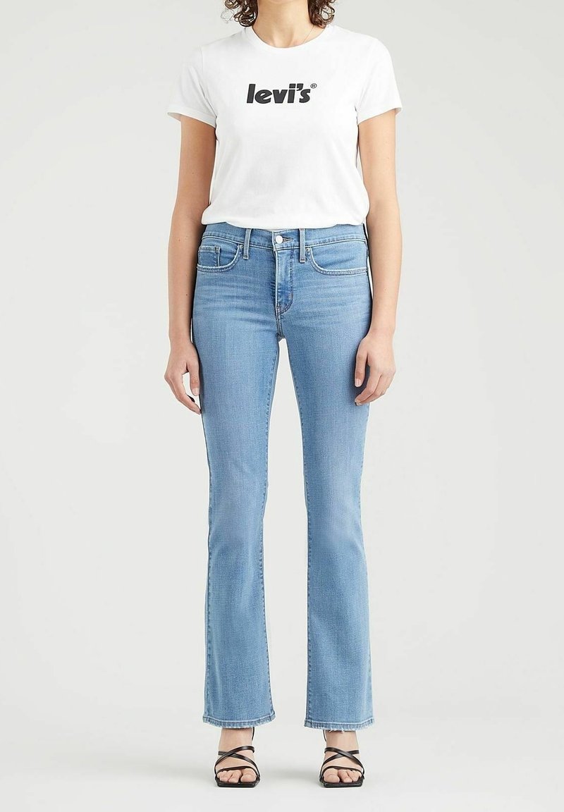 Mujer vistiendo una camiseta blanca con el logo de Levi's, jeans de cintura alta de tiro acampanado en azul claro, y sandalias de tacón negro con tiras, de pie sobre un fondo blanco.