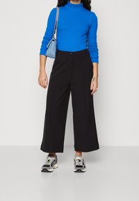 Svarta vida byxor i kombination med en blå långärmad polotröja, accessoriserat med en ljusblå handväska och grå sneakers.