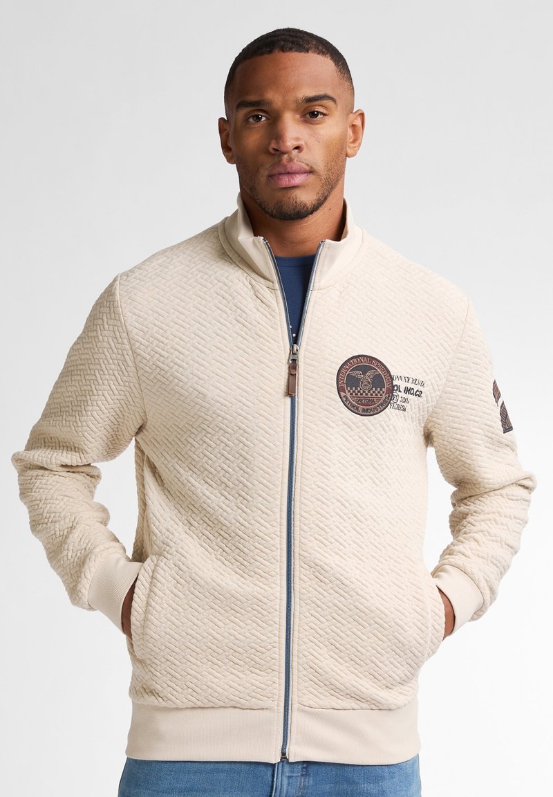 Beige tekstureret jakke med lynlås og en herringbone mønster, der har et rundt logo patch og sidelommer. Ribbestrikkede manchetter og kant.