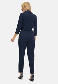 Tuta blu navy con maniche lunghe e vita regolabile. Realizzata in tessuto morbido e elasticizzato. Presenta un colletto e gambe affusolate. Indossata con tacchi alti.