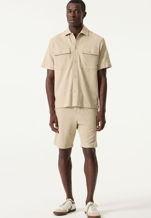 Beiges kurzärmeliges Hemd mit zwei Brusttaschen, kombiniert mit passenden Shorts. Beide Teile sind aus strukturiertem Stoff gefertigt und haben eine lockere Passform.