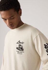 Hosszú ujjú krémszínű pulóver, amelyen egy fekete grafika látható egy teherautóról és a "Levi's" feliratról az elején; puha anyag, laza szabású.