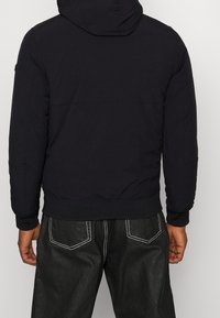 Sort hættejakke med polstret design, der har ribstrikkede manchetter og kant, kombineret med sorte teksturerede bukser med hvide syningsdetaljer.