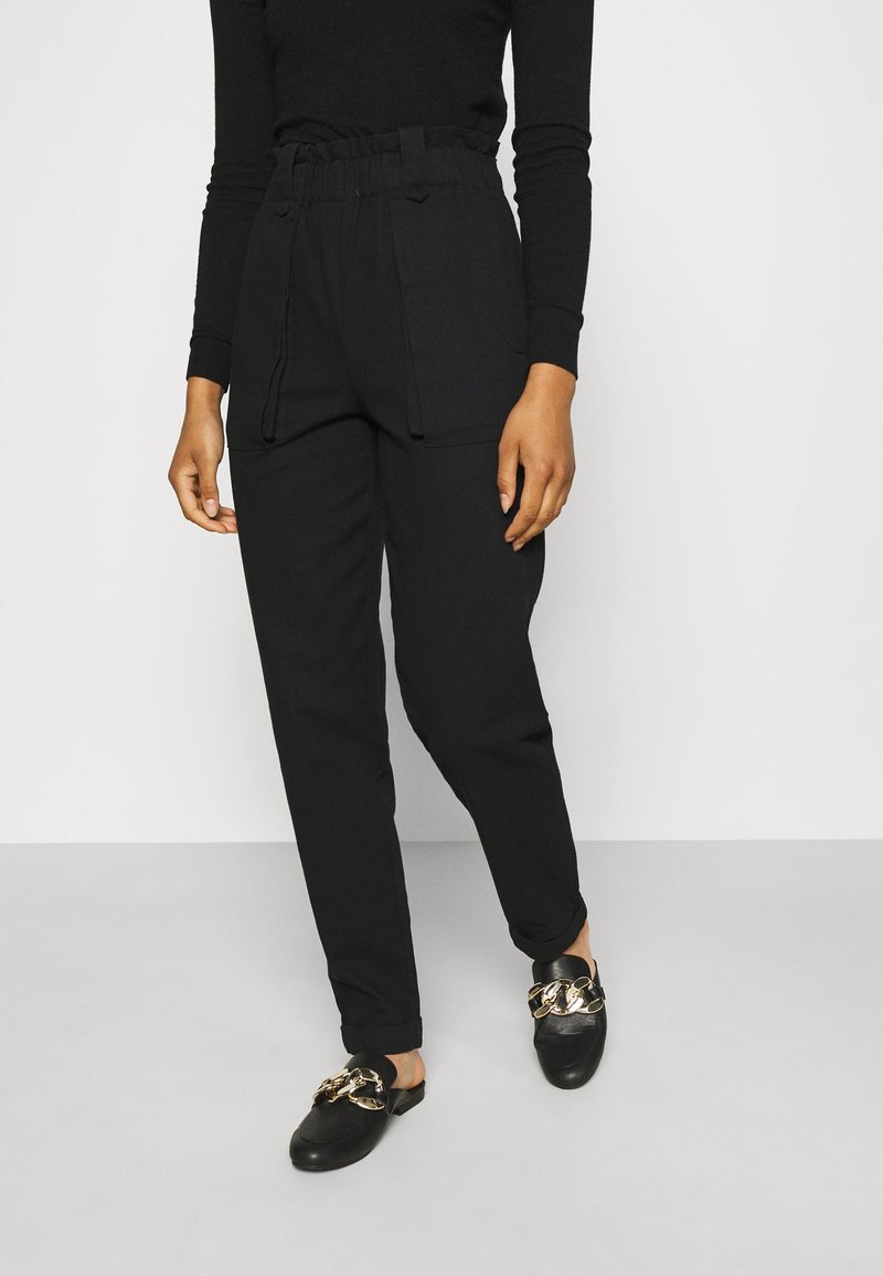 Černé kalhoty s vysokým pasem a elastickým pasem, předními kapsami a volným střihem, spárovány s černými nazouvacími botami se zlatými akcenty.