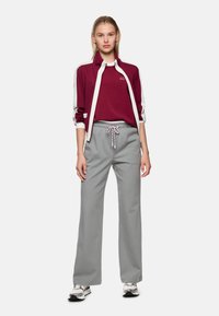 Bordeauxroter Reißverschluss-Jacke mit weißen Akzenten; kombiniert mit grauen, gestreiften Hosen mit weitem Bein und Kordelzug in der Taille. Legeres Schuhwerk rundet den Look ab.
