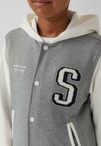 Veste à capuche grise avec des manches blanches, ornée d'une lettre "S" en tricot bleu marine. Comprend des boutons et un texte brodé sur la poitrine.