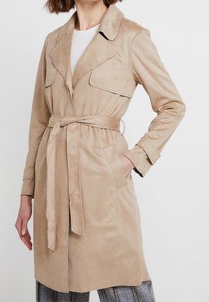 Femme portant un trench-coat beige ceinturé avec un grand col, par-dessus un haut blanc et un pantalon à carreaux, la main gauche dans la poche du manteau.