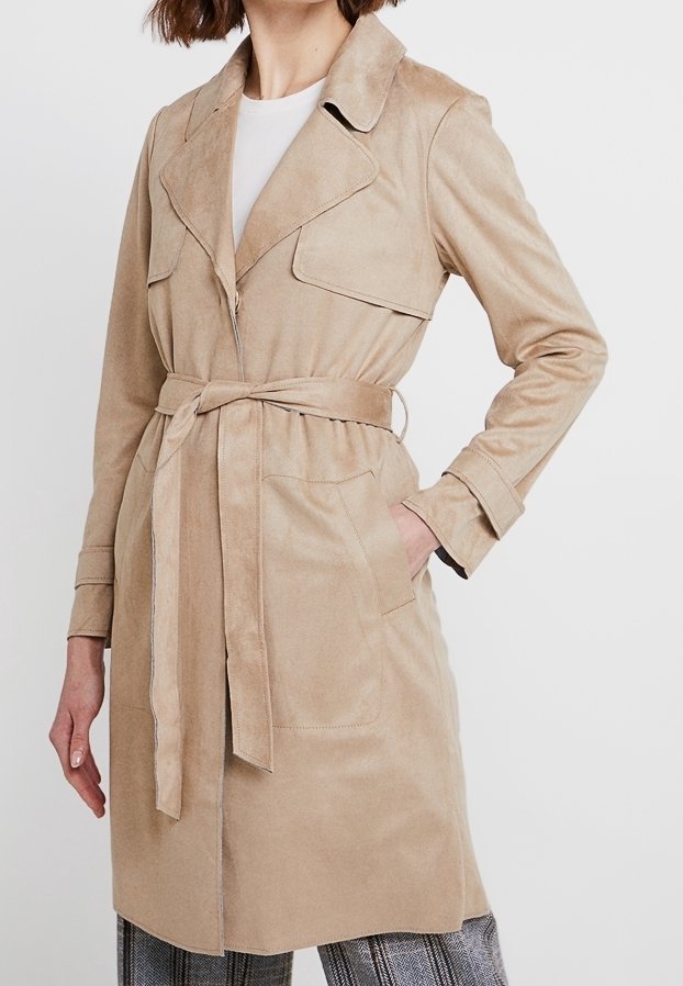 Femme portant un trench-coat beige ceinturé avec un grand col, par-dessus un haut blanc et un pantalon à carreaux, la main gauche dans la poche du manteau.