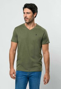 Alyvuogių žalia minkšta medvilninė marškinėliai su v formos iškirpte ir trumpomis rankovėmis. Ant krūtinės yra nedidelis tamsiai mėlynas logotipas, derinamas su mėlynais džinsais.