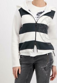 Zippar hoodieväst i mörk teal och vita ränder, texturerat stickat material, kombinerad med en vit t-shirt. Mörkgrå jeans med subtila dekorationer.