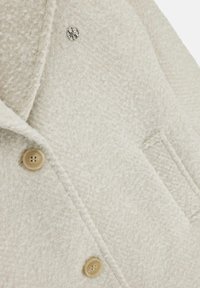 Cappotto bianco testurizzato con motivo a spiga di pesce, due bottoni e un'emblema argentato sul petto. Tessuto morbido con un design su misura.