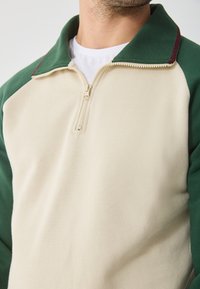 Sudadera con cremallera en color beige y verde, con cuello, que presenta un tejido texturizado y cierre de cremallera, mostrada parcialmente en la imagen.