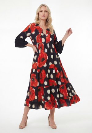 Femme blonde portant une robe longue noire ornée d'un grand motif coquelicot rouge et de pois blancs, debout avec une main sur la hanche et des talons nus.