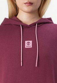 Burgundy huvtröja med en rosa logotagg och matchande rosa dragsnören. Slät bomullsstruktur, minimalistisk design och klassisk passform.