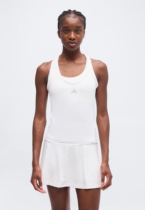 Femme aux cheveux tressés portant une robe de sport blanche sans manches avec un petit logo sur la poitrine, debout devant un fond clair uni.
