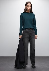 Maglione in lana turchese con scollatura a barca, abbinato a jeans neri a gamba larga. Il modello tiene un giubbotto nero e indossa stivali neri con tacco alto.