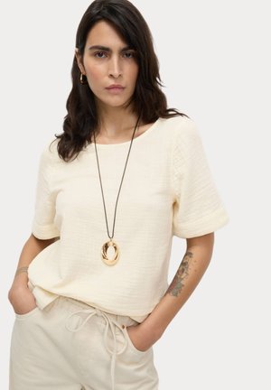 Top corto a maniche corte di colore crema, con una vestibilità comoda e texture. Abbinato a pantaloni beige e a una lunga collana con un pendente metallico.