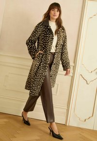 Barna mintás kabát fekete gallérral, hosszú ujjakkal és zsebekkel, barna bőr nadrággal és fekete magassarkú cipővel párosítva.