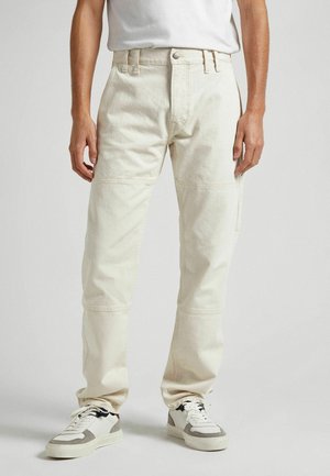 Mies, joka käyttää luonnonvalkoisia suoria housuja saumat yksityiskohtina, valkoisia ja harmaita tennareita sekä valkoista paidan sisään pantuja, seisoo vaaleaa taustaa vasten.