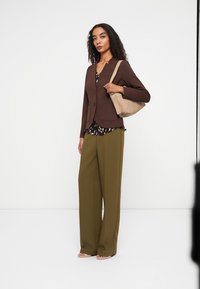 Brauner, taillierter Blazer mit zwei vorderen Taschen über einer bedruckten Bluse, kombiniert mit olivgrünen, weit geschnittenen Hosen und einer beigen Schultertasche.