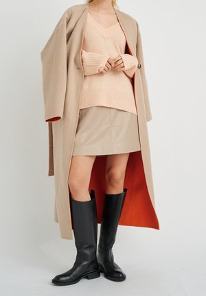Kobieta w beżowym długim płaszczu z pomarańczową podszewką, brzoskwiniowym swetrze z dekoltem w serek, beżowej spódnicy i czarnych kozakach za kolano, stoi na jednolitym tle.