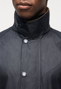 Άνδρας που φοράει σκούρο σακάκι με ψηλό γιακά και δύο εμφανή στρογγυλά ασημί κουμπιά, κοντινό πλάνο που δείχνει μόνο τα χείλη και το κάτω μέρος του προσώπου.