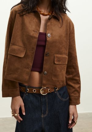 Persoon draagt een cropped bruine suède jas, een bordeaux crop top, donkere spijkerbroek met een bruine riem, een amberkleurige ketting en meerdere ringen.