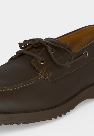 Chaussure bateau en cuir marron foncé avec couture moc toe, laçage à l'avant, œillets en laiton et semelle en caoutchouc sur fond uni.