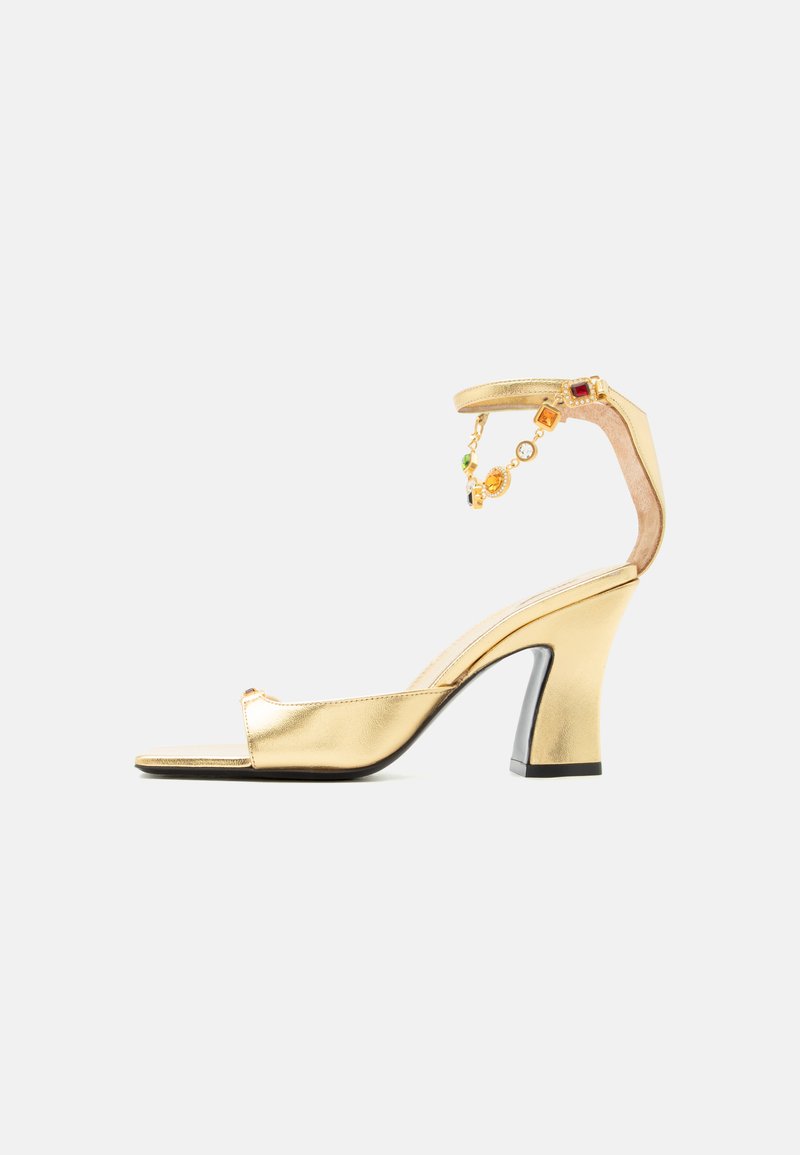 Zlatá sandála na vysokom podpätku s remienkom okolo členka, zdobená farebnými kamienkovými príveskami, otvorenou špičkou a blokovým podpätkom na bielom pozadí.