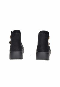 Stivaletti ankle in suede neri con piattaforma, impreziositi da fibbie floreali dorate, visti da dietro su uno sfondo bianco.
