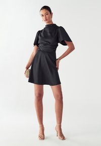 Crna saten mini haljina s kratkim puf rukavima, uskim korpusom i glatkim naborima. Uparena s bež sandalama s trakama i malom torbicom.