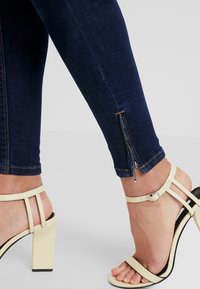 Jeans skinny azul oscuro con detalle de cremallera en el tobillo, combinados con unas sandalias de tacón alto beige que cuentan con tiras finas y un tacón cuadrado.