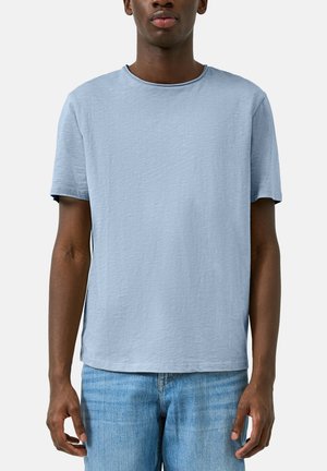 Mann trägt hellblaues kurzärmeliges T-Shirt und blaue Jeans, steht vor einem schlichten weißen Hintergrund.